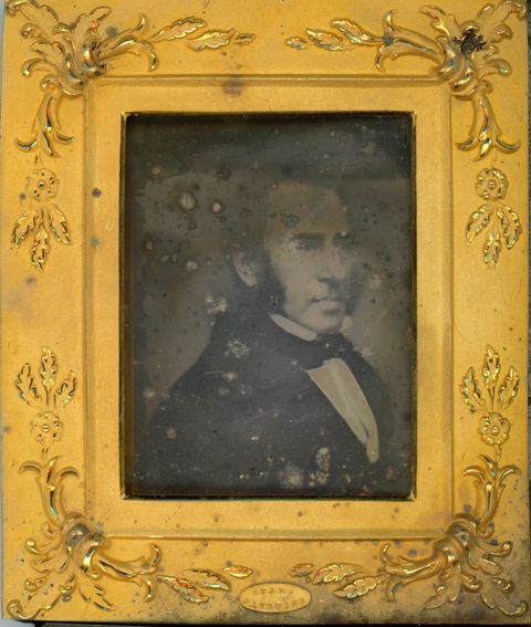 Daguerreotype. A pale-skinned man with bushy mutton chops looks off to our right. He is framed in the rectangular opening of a gold mat, which is decorated at the corners with flowers and leafy designs in low relief.