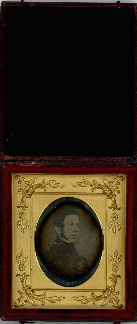 Daguerreotype in an open case. A pale-skinned man with long mutton chops looks off to our right. He is framed in the oval opening of a gold mat, which is decorated at the corners with flowers and leafy designs in low relief. The case flips up, and the cover is lined with dark red fabric.