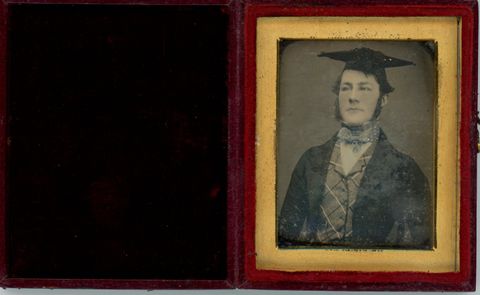 Daguerreotype in an open case. A pale-skinned young man wearing a mortar board cap looks stiffly off to our left. He wears a vest, jacket, and a cravat coloured light blue. The photograph is in a gold frame in a red case, which opens like a book. The cover is lined with black fabric.