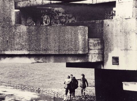 Architecture. In this black and white photograph, concrete planes and ledges make up a bewildering space. Three children stand by water at the concrete edge of an aqueduct.