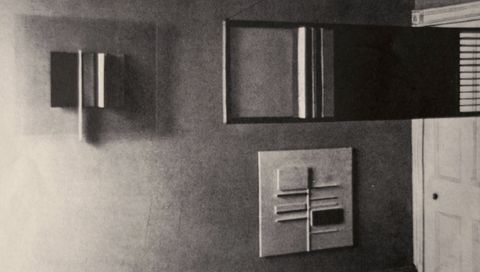 Installation photograph. Abstract works of art comprising stacked panels in rectilinear geometric designs are arranged on and in front of a wall in this black and white photograph.