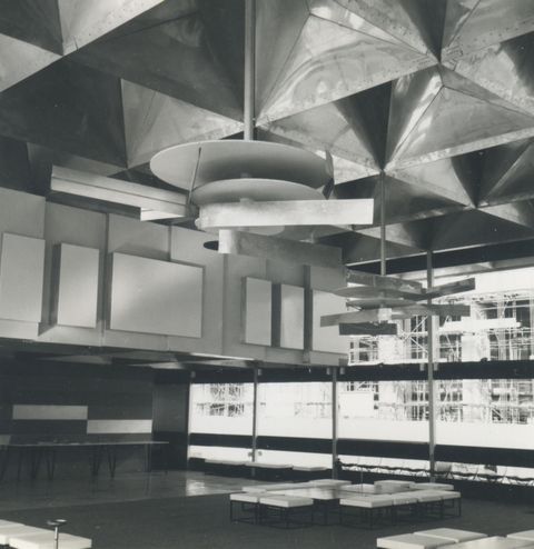 Installation photograph. Black and white photograph of a window-lined room with deep, angular coffers in the ceiling above sleek-lined furniture. Two abstract sculptures hanging from the coffers are made up of flat disks and long strips of metal.