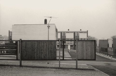 Architecture. Panels of vertically oriented fencing and horizontal boards are against blocky buildings with rectangular windows in this black and white photograph.