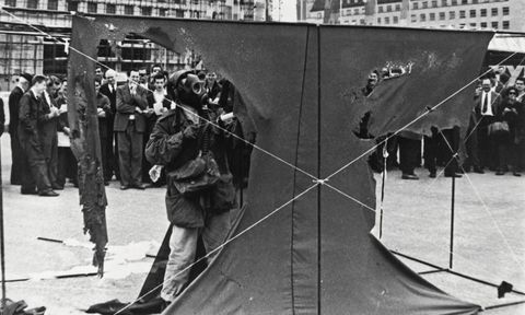 Performance art. A piece of dark cloth the size of a single car garage is stretched across two rails, one above and one below, in this black and white photograph. From the far side of the cloth, we face the artist, who wears a gas mask and military jacket as he sprays acid onto the cloth. Large swaths have been chewed away, leaving an inwards-curving, wide, T shape of fabric behind.