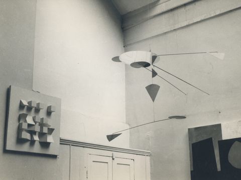 Installation photograph. In this black and white photograph, sculptural works are displayed a high-ceilinged room. To our left is a flat white square from which sixteen cubes, triangles, and other polygonal blocks project in a grid. A mobile with fine arms that suspend flat geometric shapes hangs from the ceiling. The edge of another abstract work is visible in the lower right corner of this view.