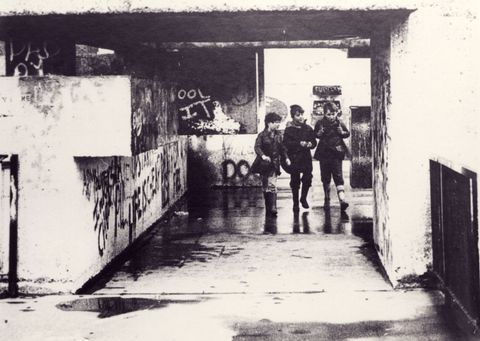 Architecture. Three boys walk towards us through a squared concrete opening lined with graffiti in this black and white photograph.