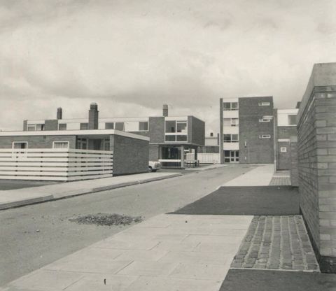Architecture. Black and white photograph of blocky residential buildings and horizontal fencing. The bildings are between one and four stories high. A street that recedes across the image and its sidewalks are empty.