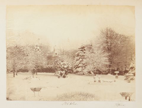 Black and white photograph. A snow-covered lawn leads back to trees lining a lane. A steeple-like tower is nestled in the trees along the horizon. Handwriting below reads “Stables” and “Pitfour”.