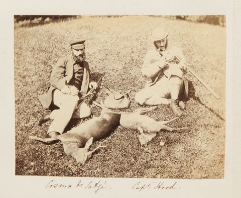 Black and white photograph. Two pale-skinned men recline in the grass above two dead deer. The men have rifles and wear caps and suits with vests. The man to our right has a clay pipe in one corner of his mouth. The men’s names are written in pen below.