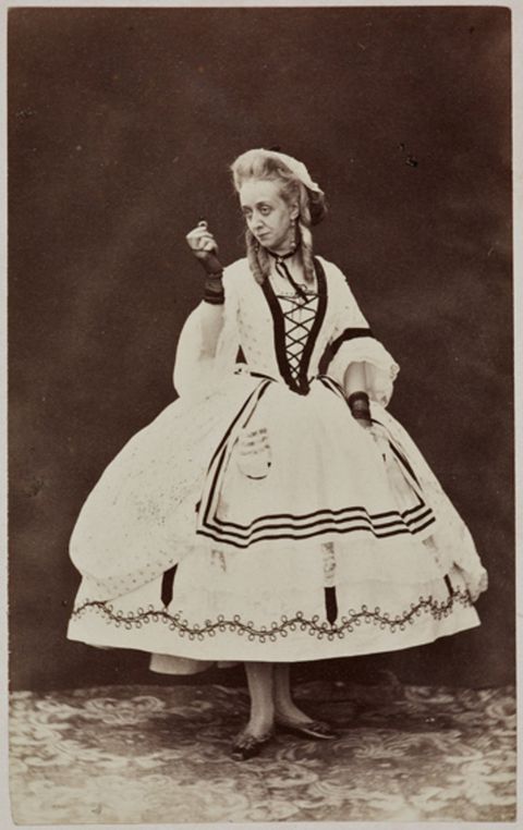 Black and white photograph. A pale-skinned woman wears a white dress edged with black lines or designs. The skirt flares out from the hips and ends above the ankles. She holds up a ring with her right hand, to our left, and looks in that direction against a dark background.