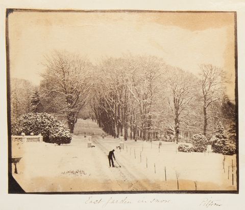 Black and white photograph. We look slightly down onto a worker in black silhouette against the white ground clearing snow from a lane. Snow-laden trees line the drive and enclose the space. The title is written in pen below along with the location, “Pitfour”.