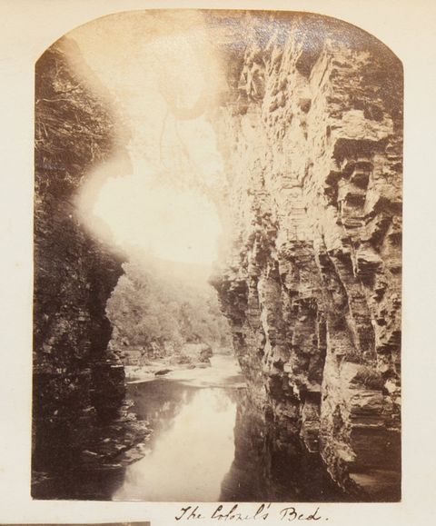 Black and white photograph. Sheer, rocky cliffs tower up both sides of this photograph. The river running between them and away from us appears to have a smooth surface. Sunlight washes out the top centre of the photograph, over a tree-lined hill. The title is written in pen below.