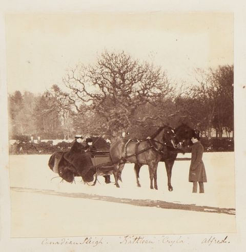Black and white photograph. Two dark horses stand ready to pull a sleigh occupied by two women. A man wearing a long coat stands at the horses’ heads. Leafless trees are lacy against a blank sky over an expanse of white, snowy lawn. The title and peoples’ names are written in pen below.