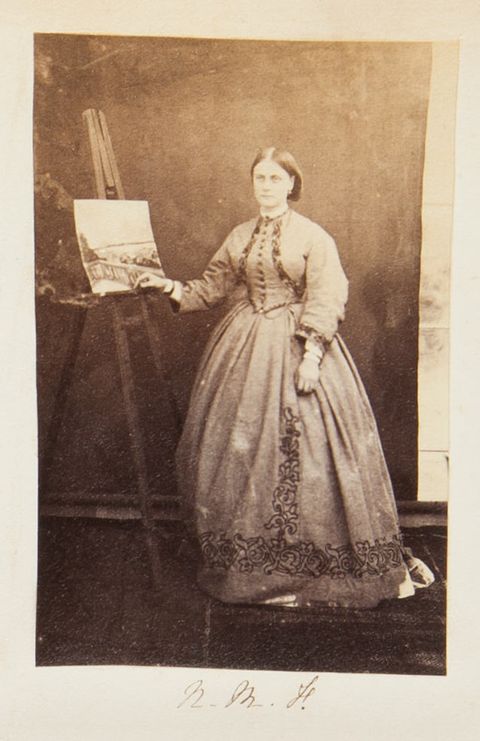 Black and white photograph. A pale-skinned woman wearing a long-sleeved dress with a full, wide skirt stands with one hand resting on the edge of an easel, which holds a painting.