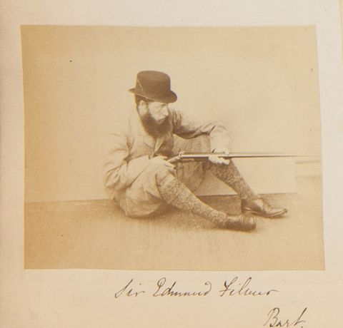 Black and white photograph. A man with pale skin and a full, bushy beard sits on the ground, knees bent so he can steady a rifle. He faces our right almost in profile. His name is written in pen below.