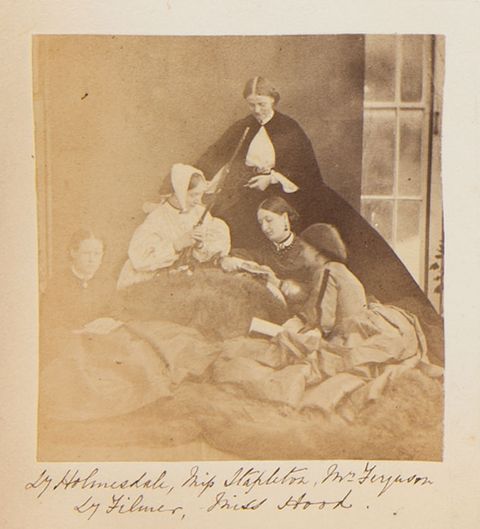 Black and white photograph. An enormous fur blanket is draped over one woman’s lap and across the ground. Three more women sit on the fur, reading books. The woman with the fur over her legs holds a long-barrelled gun angled upwards. A woman in black is at the apex. The women are identified in handwriting below.