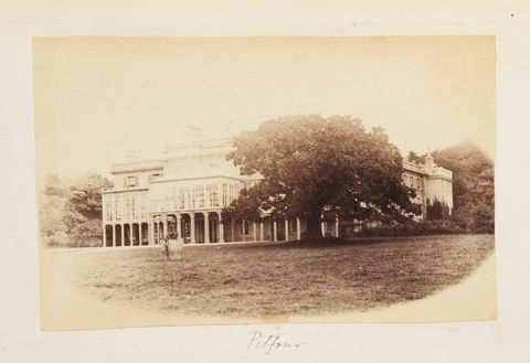 Black and white photograph. A three-storey mansion sits behind a sprawling tree. The lowest level is lined with columns to create a long covered walkway or porch.