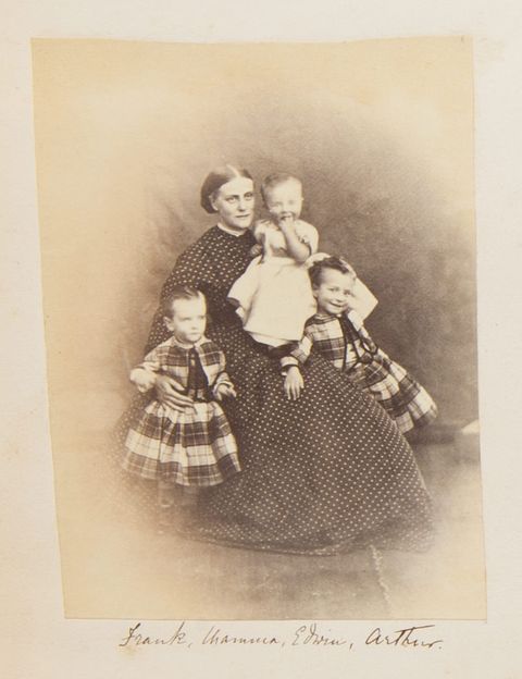 Black and white photograph. A pale-skinned woman wearing a dark dress with white polka dots sits and holds a baby as two small children lean against her lap. The standing children wear plaid garments and one of the smiles at us. The baby holds one fist in their mouth and also smiles.