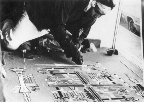 Installation. A dark-skinned man carefully drops sand from his fist to create a stylized image of people and natural forms like trees. He works on the floor, and the back end of a car can be seen out a window next to the artist.