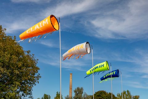 Public art. Four windsocks fill with air so a single word on each can be read. In white letters against an orange sock, the one closest to us reads “Claim”. In orange against white, the next reads “Water”. Then in blue against neon green is “Thrash” and, finally, in green against blue, “Fight”.