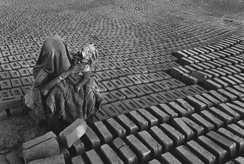 Black and white photograph. We look slightly down onto a woman sitting and holding a baby amid rows of bricks that surge and ebb with the lay of the land. The woman and baby’s faces and most of their bodies are covered with patterned cloth but their arms are dark skinned.