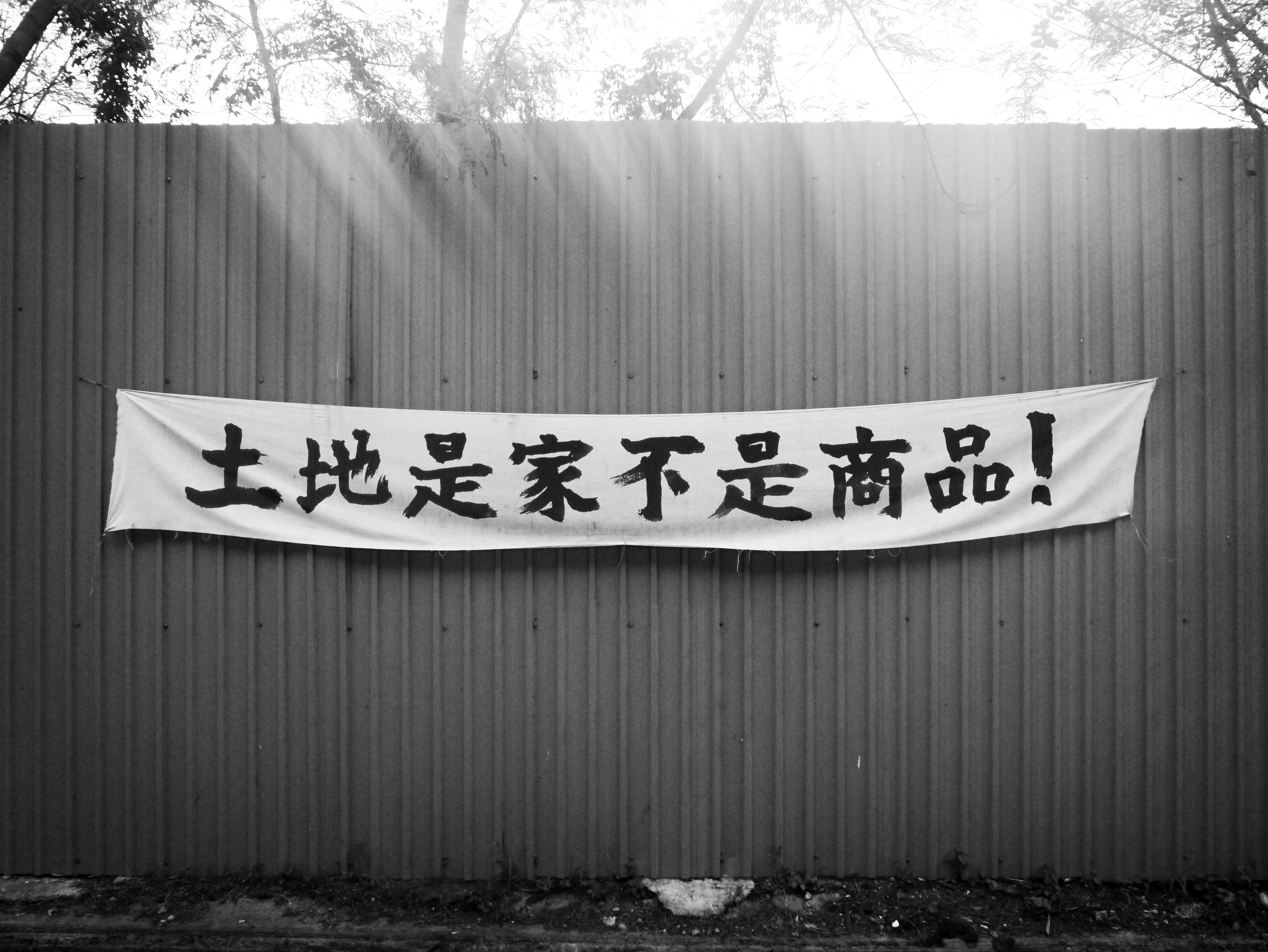 Mixed media. A black and white photograph sun streaking over the top edge of a solid fence holding a wide white banner. Chinese characters have been written across the banner.