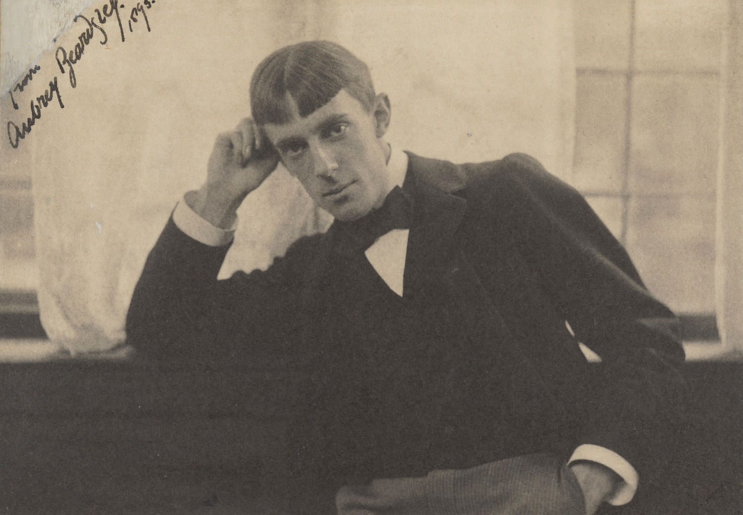 Black and white photograph. Shown from the waist up, a pale-skinned man with a long, narrow face sits with one elbow propped on a window ledge behind him. His hair is parted down the middle and cut into straight bangs across his high forehead.
