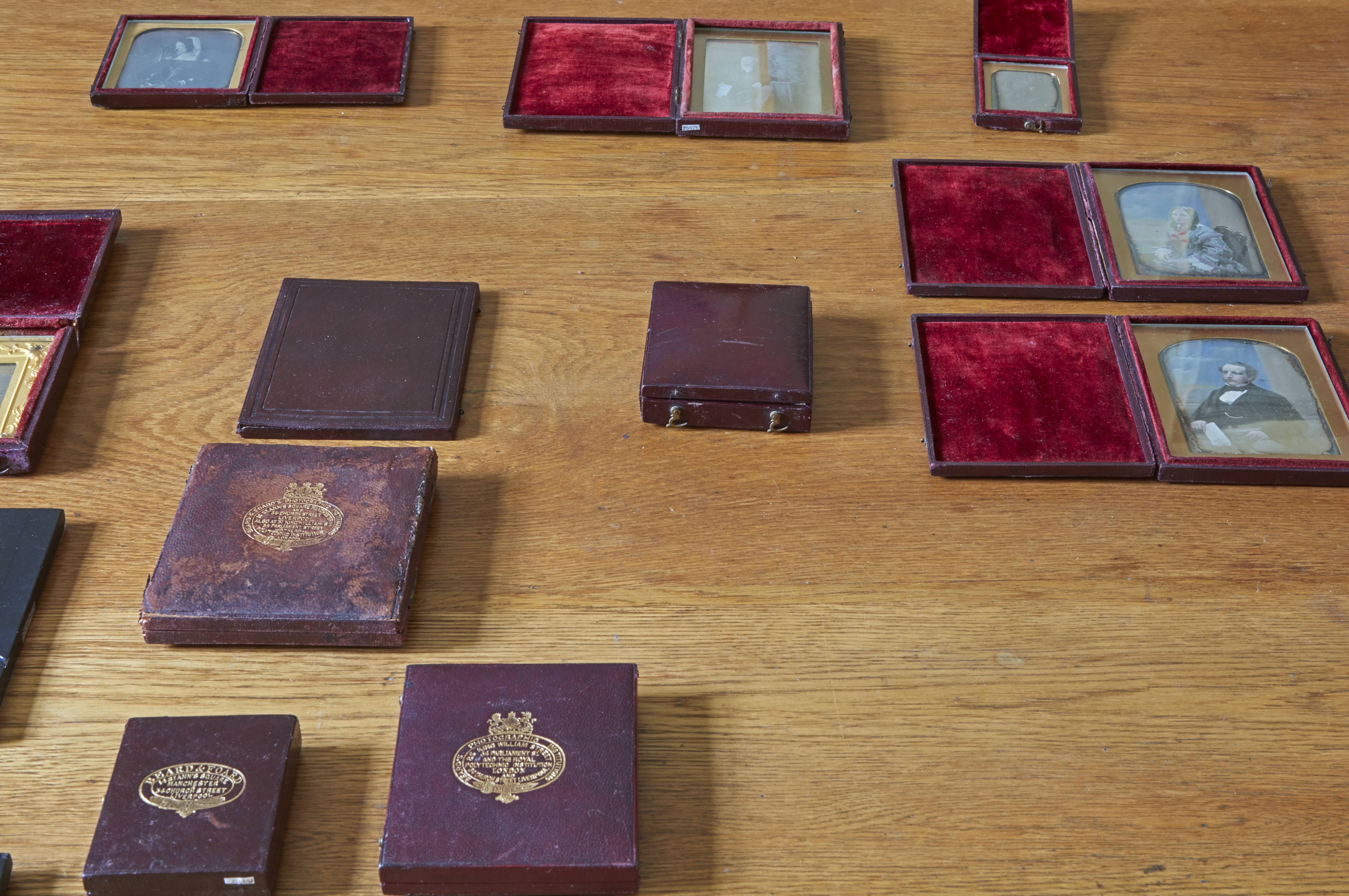 Colour photograph. Six daguerrotype portraits in fabric-lined, open cases along with five closed cases are arranged on a wooden tabletop. Light reflects off the glass fronts of the open photographs.
