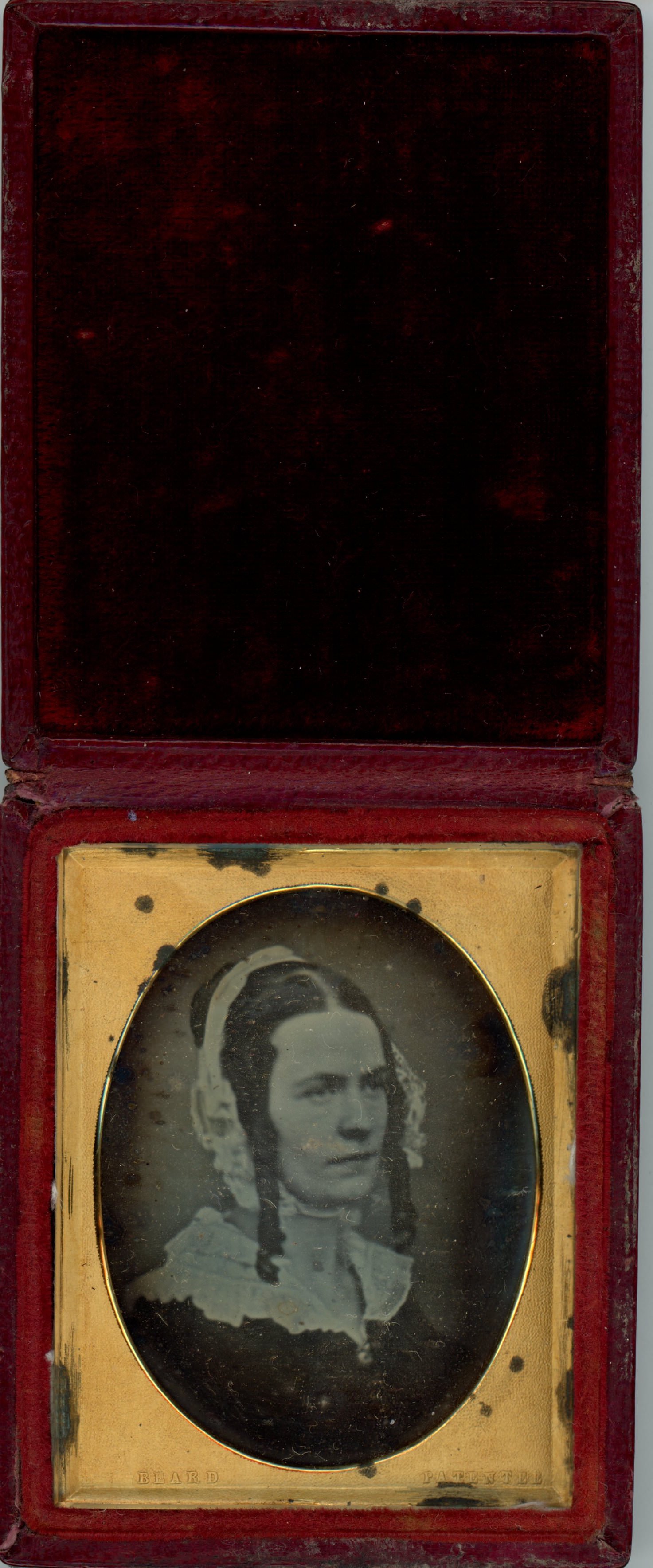 Daguerreotype in an open case. A pale-skinned woman with long, dark curls framing her face looks off to our right within the oval opening of a gold mat. The case is flipped up and lined with dark red fabric.