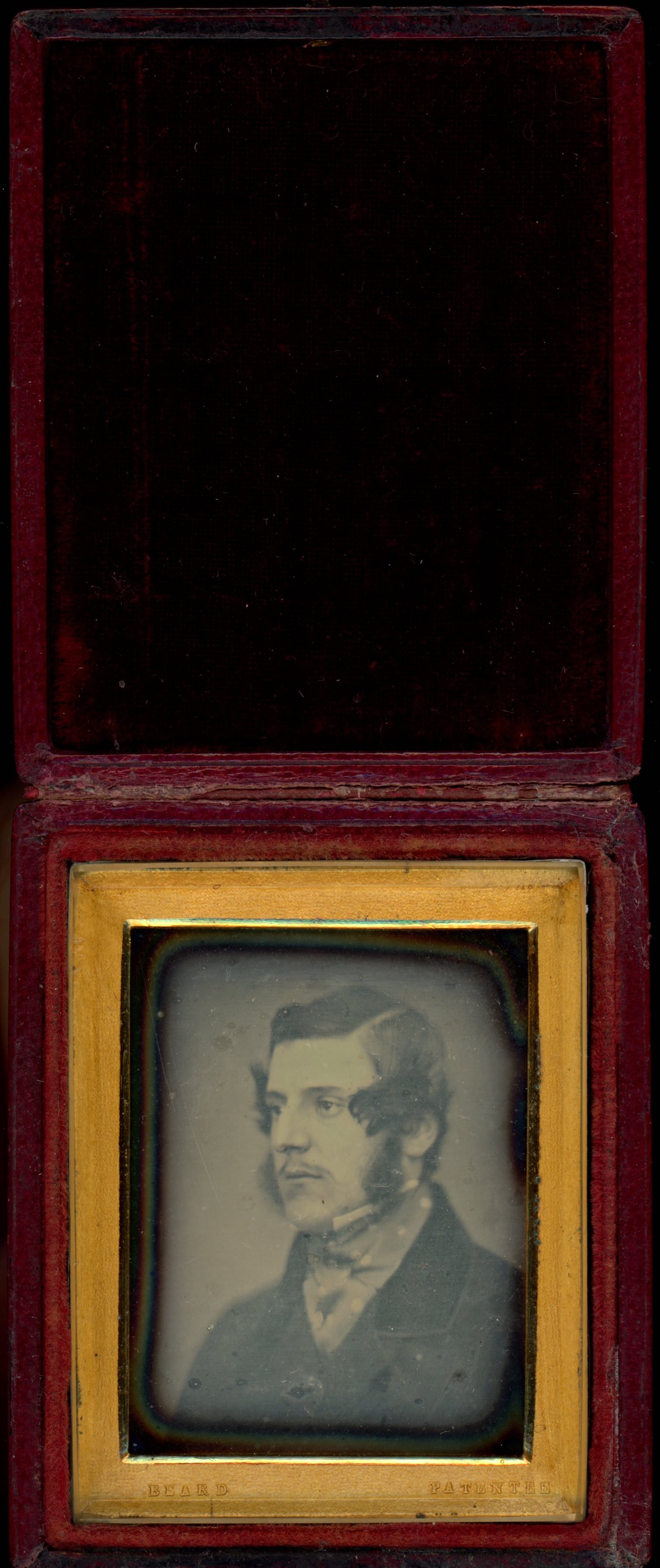 Daguerreotype in an open case. A pale-skinned man with mutton chops looks off to our left within a rectangular gold mat. The case is flipped up and lined with dark red fabric.