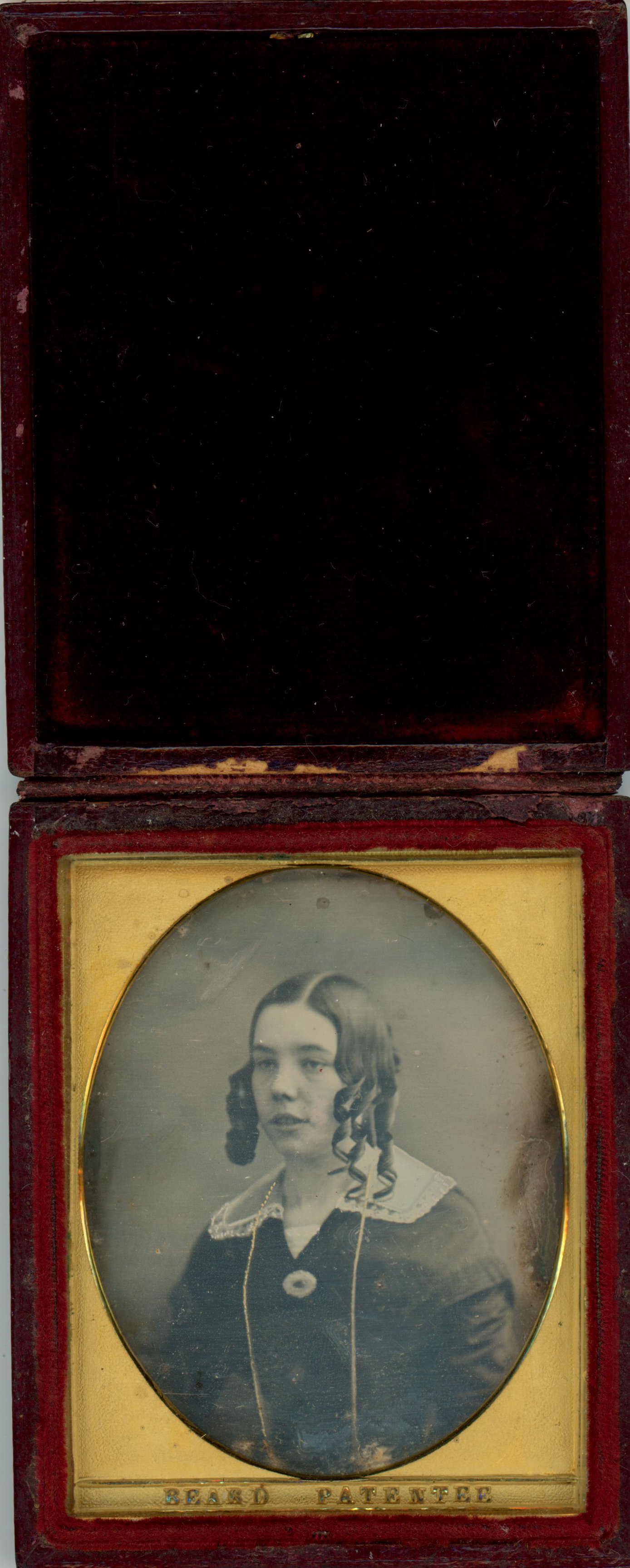 Daguerreotype in an open case. A pale-skinned woman with rows of long curls to either side of her oval face looks off to our left within an oval opening of the gold mat. The case is flipped up and lined with dark red fabric.
