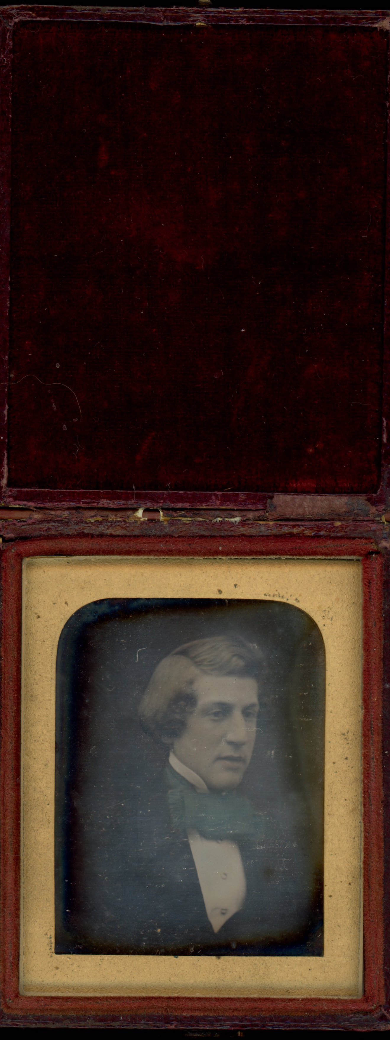 Daguerreotype in an open case. A pale-skinned, clean-shaven man looks off to our right with his thick hair combed down one side of his face and the rest over the crown. The gold mat has arched top corners. The case is flipped up and the cover lined with dark red fabric.