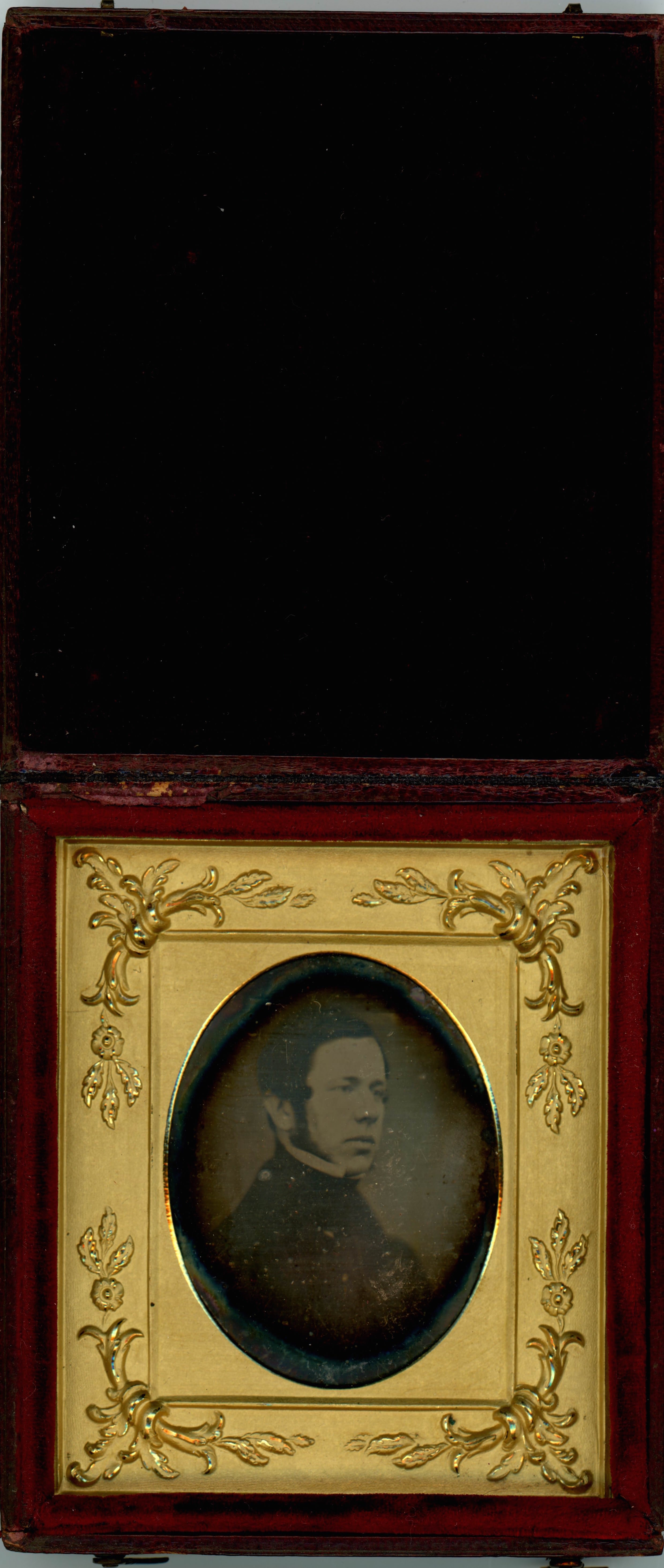 Daguerreotype in an open case. A pale-skinned man with long mutton chops looks off to our right. He is framed in the oval opening of a gold mat, which is decorated at the corners with flowers and leafy designs in low relief. The case flips up, and the cover is lined with dark red fabric.