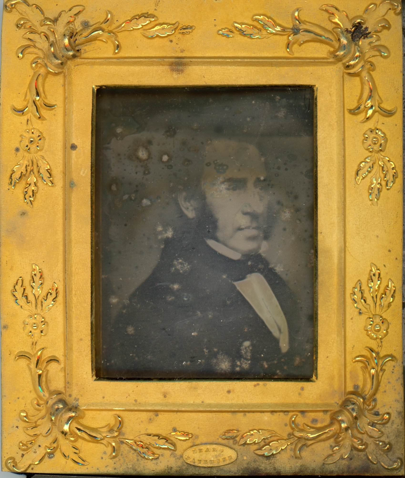 Daguerreotype. A pale-skinned man with bushy mutton chops looks off to our right. He is framed in the rectangular opening of a gold mat, which is decorated at the corners with flowers and leafy designs in low relief.