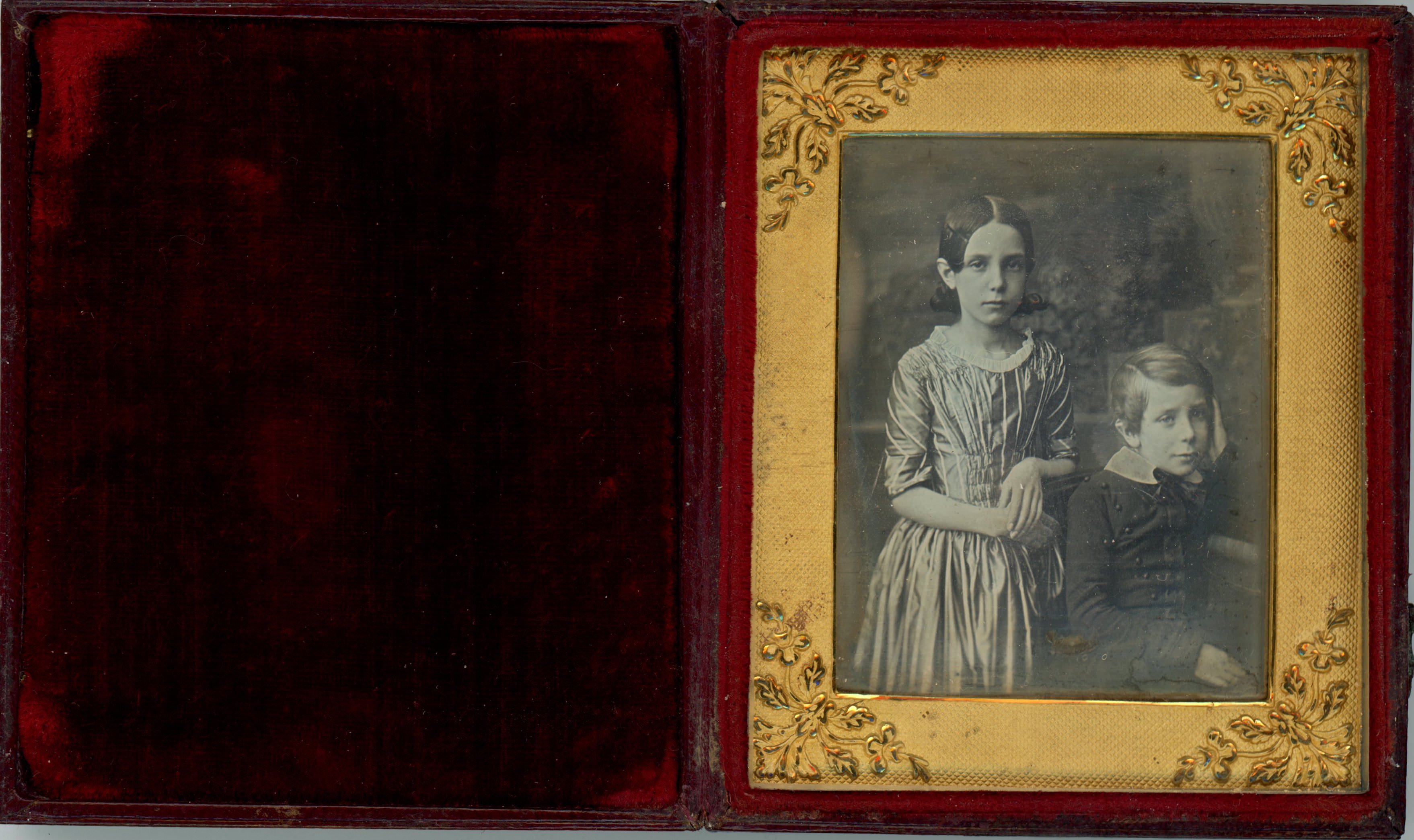 Daguerreotype in an open case. A young girl stands and a boy sits, and they look out at us. They both have pale skin and prop their left elbows, to our right, on ledges behind them. They are framed in the rectangular opening of a gold mat, which has leafy designs at the corners. The case flips up, and the cover is lined with deep red fabric.