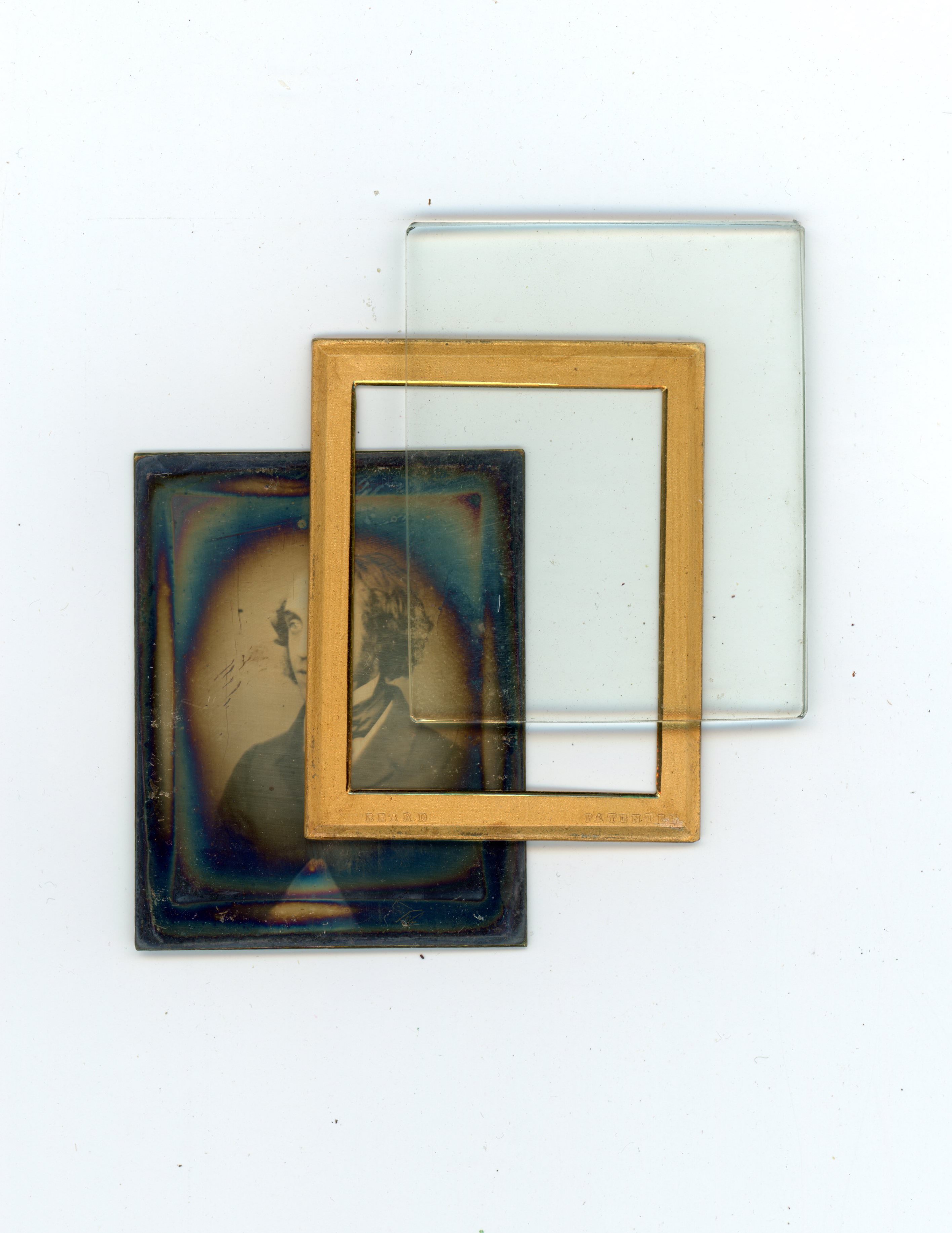 Colour photograph. Three parts of a framed daguerreotype are shown. The photograph has iridescent rainbow edges and is accompanied by a gold mat and a piece of glass to show how the photograph can be protected in a case.