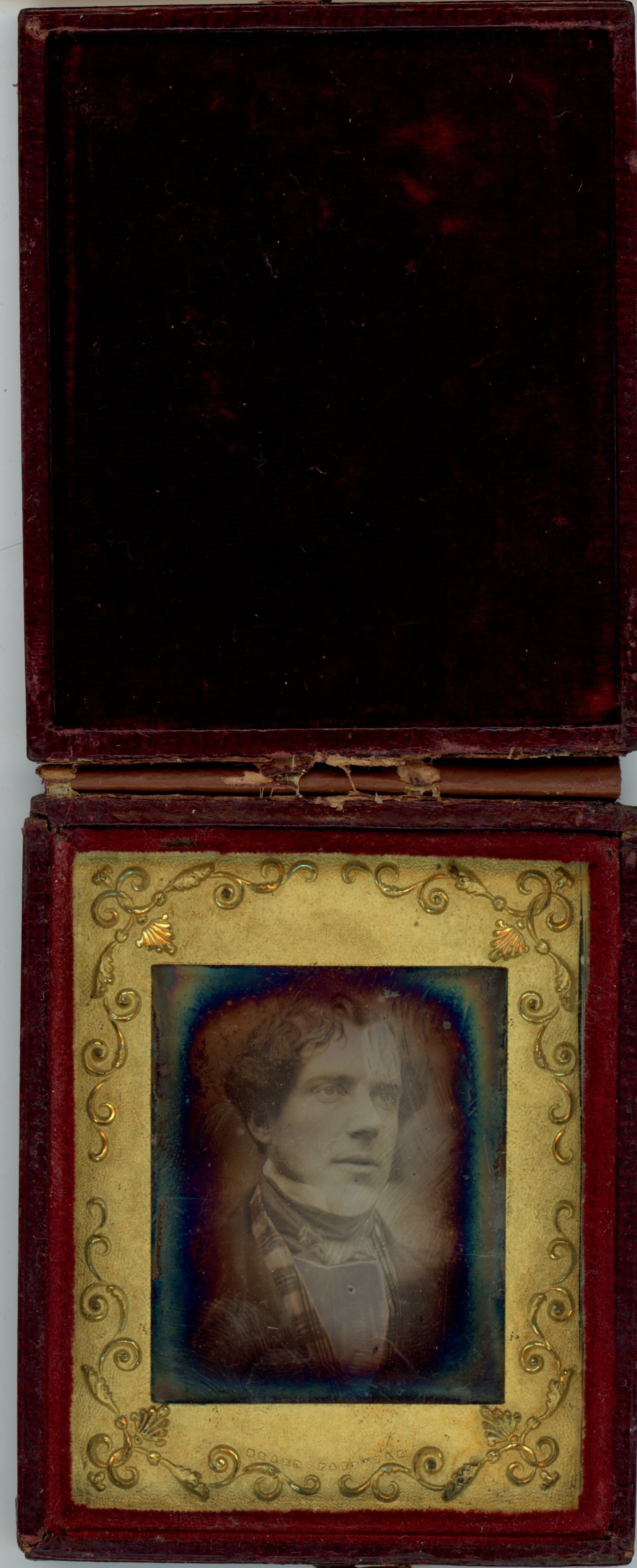 Daguerreotype in an open case. A pale-skinned man with dark hair and short mutton chops looks off to our right. The edges of the photograph are rainbow coloured, like oil on water. He is framed in the rectangular opening of a gold mat, which is decorated with scrolling vines. The lid of the case is flipped up, and the underside is lined with dark red fabric.