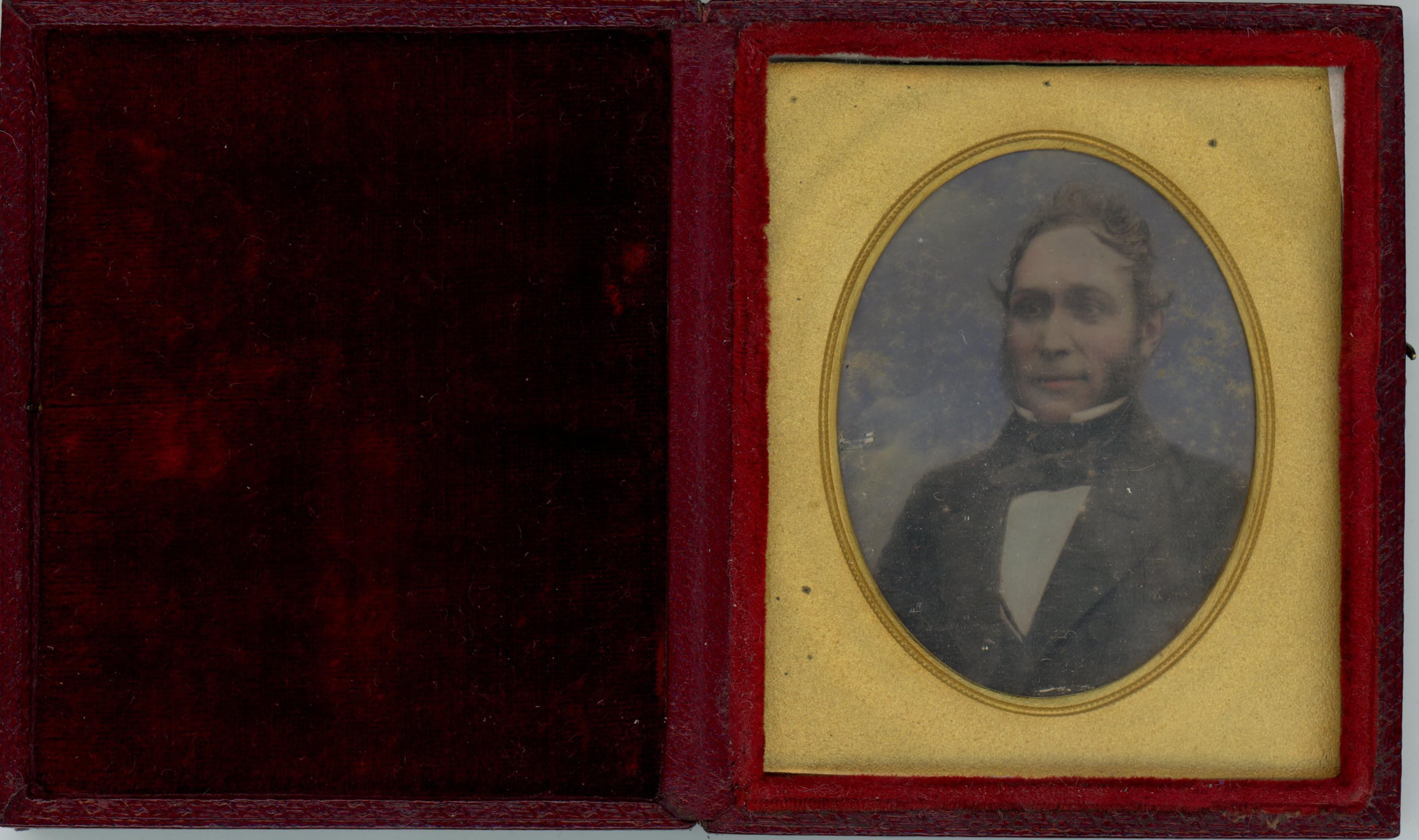 Daguerreotype in an open case. A pale-skinned man looks off to our left. His cheeks and lips have been tinted pink, and the background tinted blue. He is framed in the oval opening of a gold mat. The lid of red case is flipped open to our left and is lined with dark red cloth.