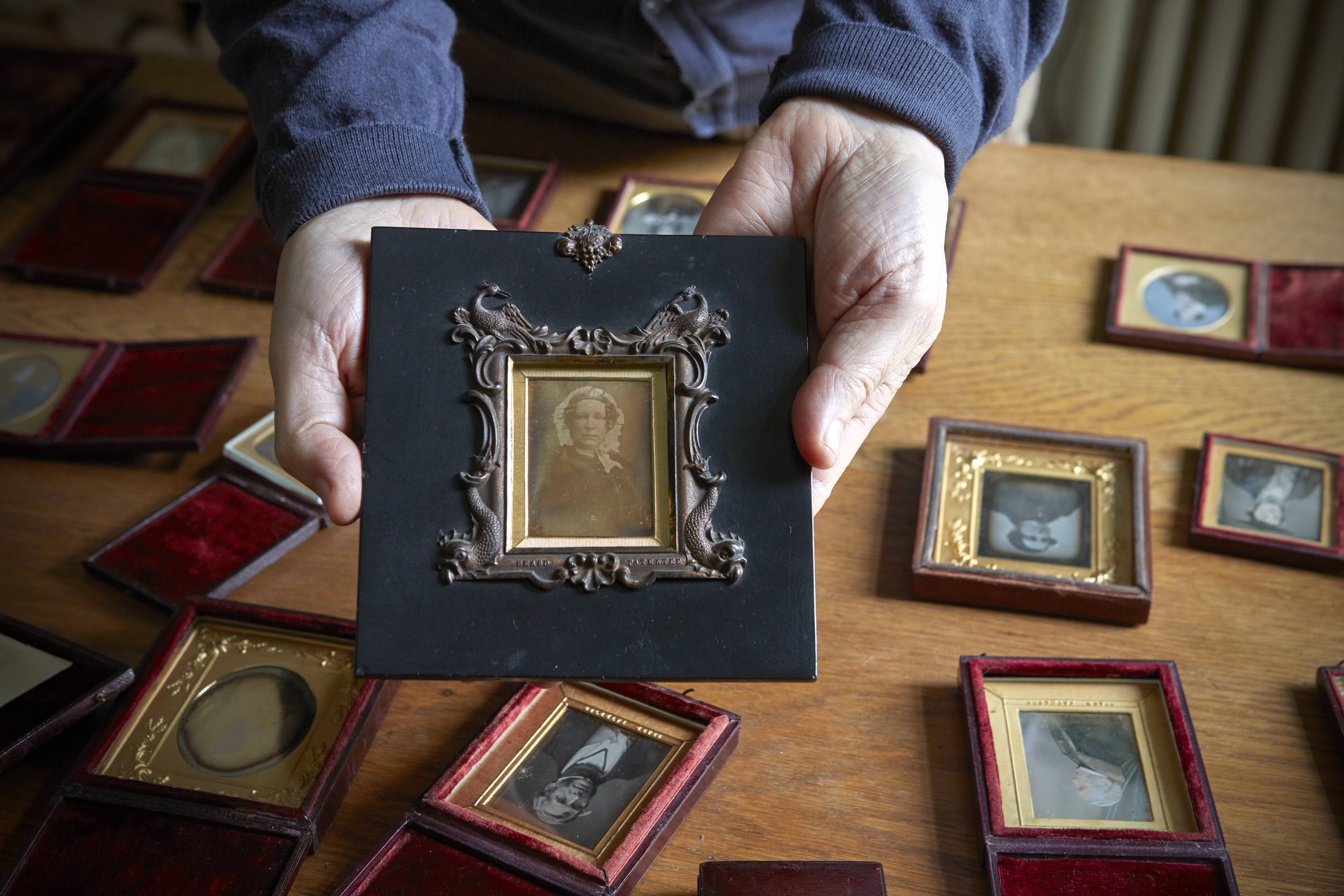 Colour photograph. Two hands hold a framed daguerreotype portrait of a woman wearing a lacy bonnet. The frame around the gold mat is raised in a design of birds and stylized fish. More daguerreotypes are laid out on a tabletop behind the hands.