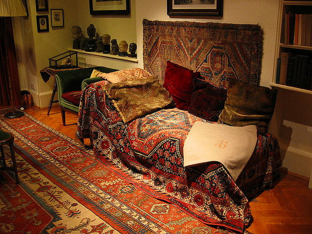Colour photograph. A lounging couch is piled with patterned fabrics. Velvet cushions lean against the wall along one long side. The room has a patterned carpet on the floor and another on the wall behind the sofa. Sculpted heads and books line shelves around the couch.