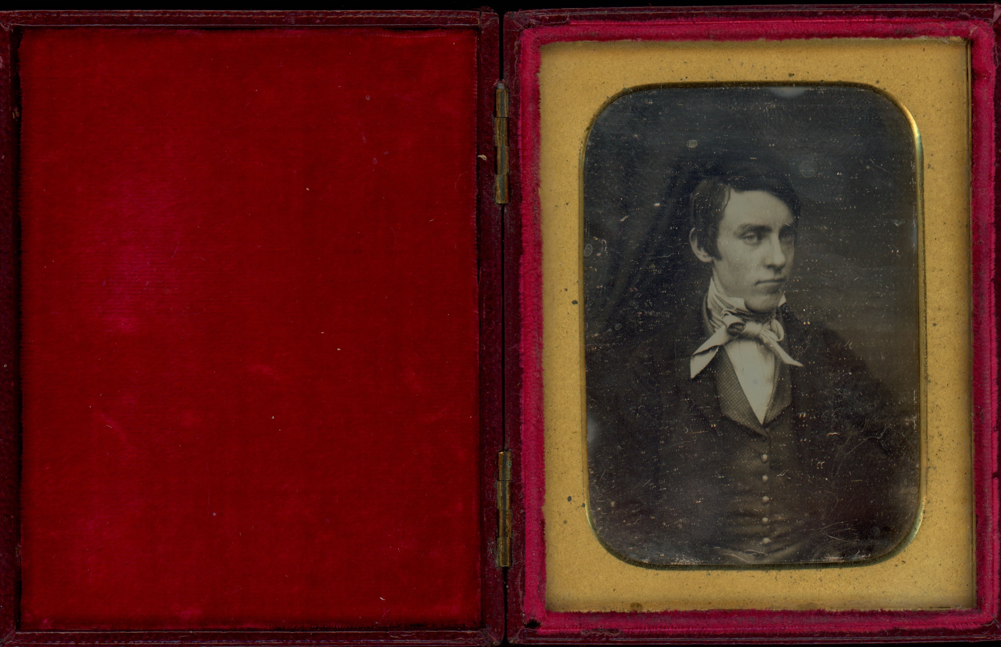 Daguerreotype in an open case. A pale-skinned, clean-shaven young man’s face is angled to our right, but he looks at us under heavy brows, his chin slightly tucked. The rectangular mat has curved edges. The crimson-red case is flipped open to our left.