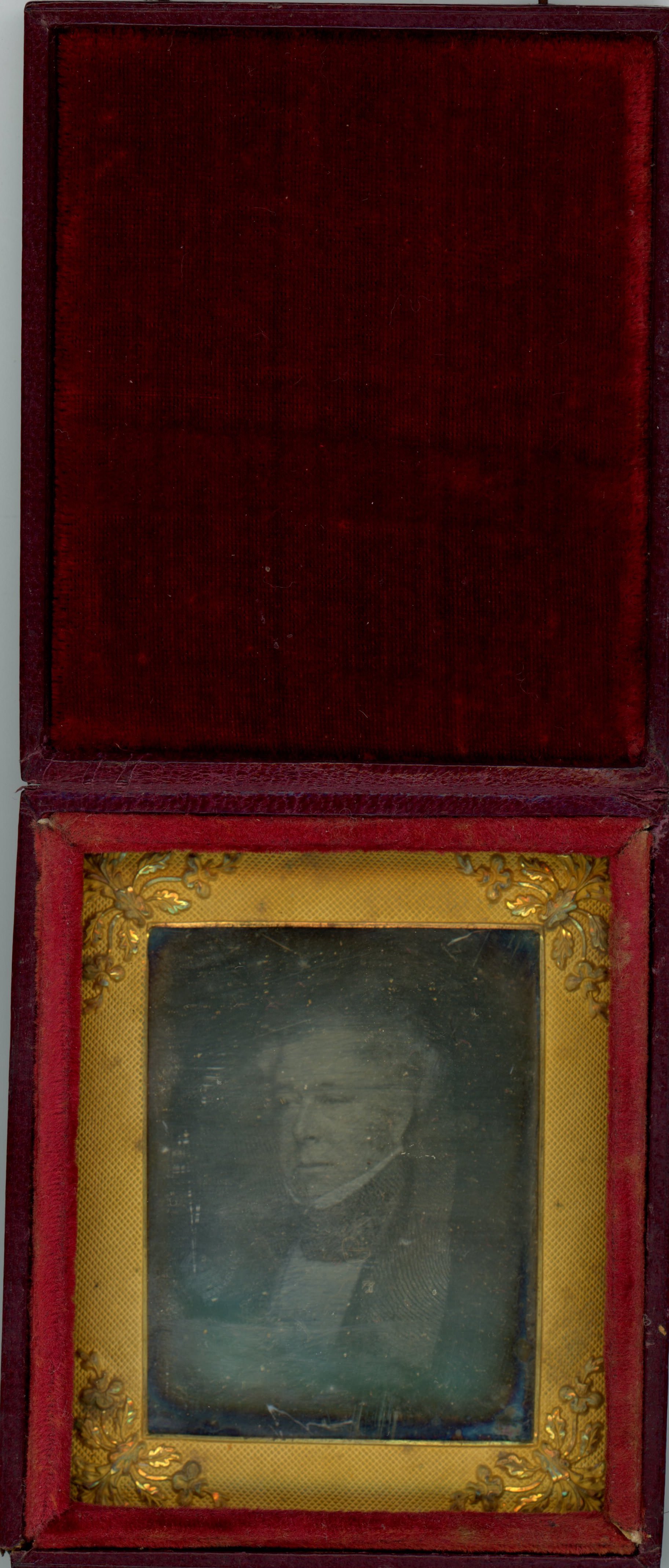 Daguerreotype in an open case. The ghostly image of a clean-shaven man with white or grey hair looks off to our left. The rectangular mat has leafy designs at the corners. The lid of the case is flipped up, the inside lined with dark red velvet.