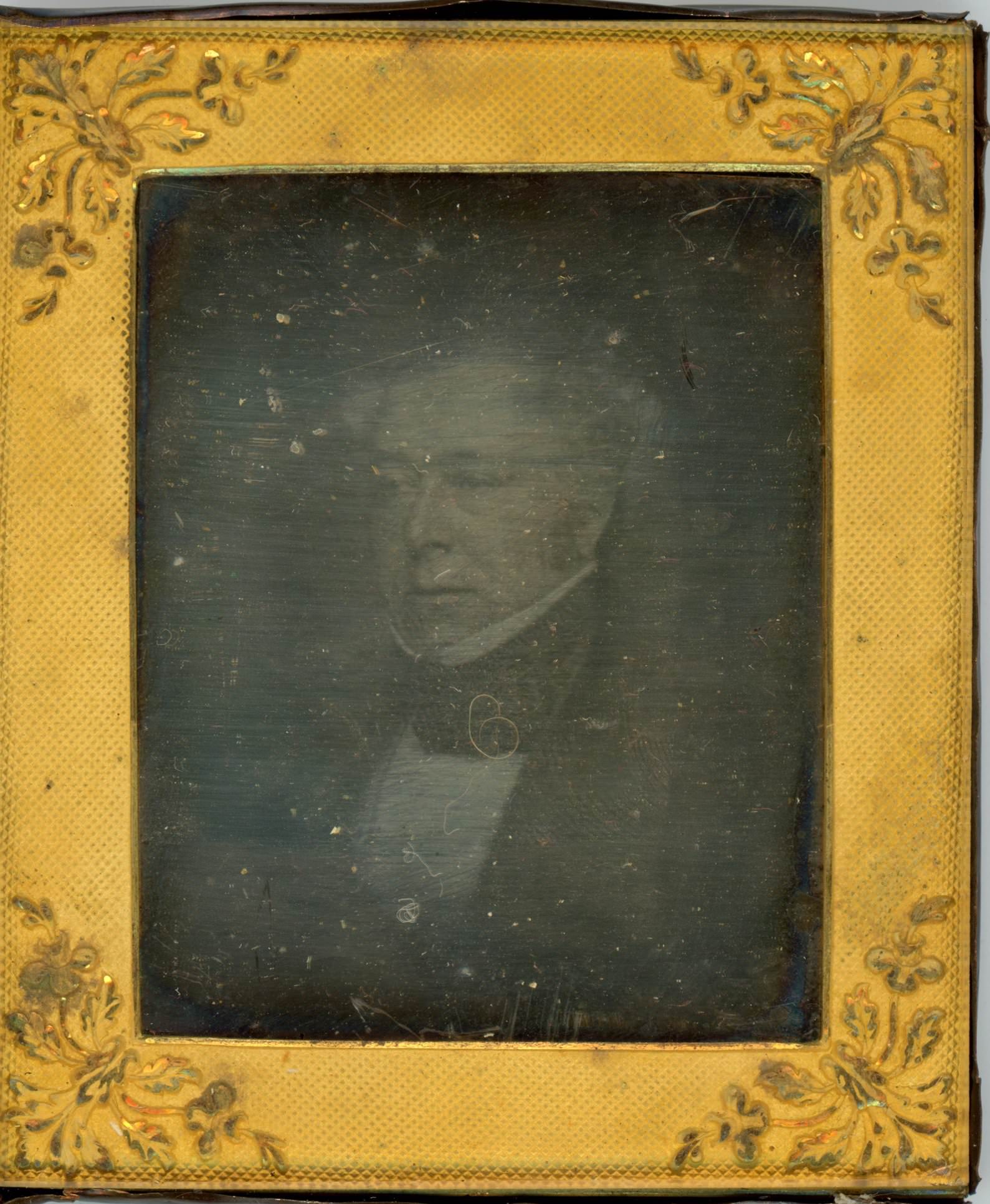 Daguerreotype. The ghostly image of a clean-shaven man with white or grey hair looks off to our left. The gold rectangular mat has leafy designs at the corners.