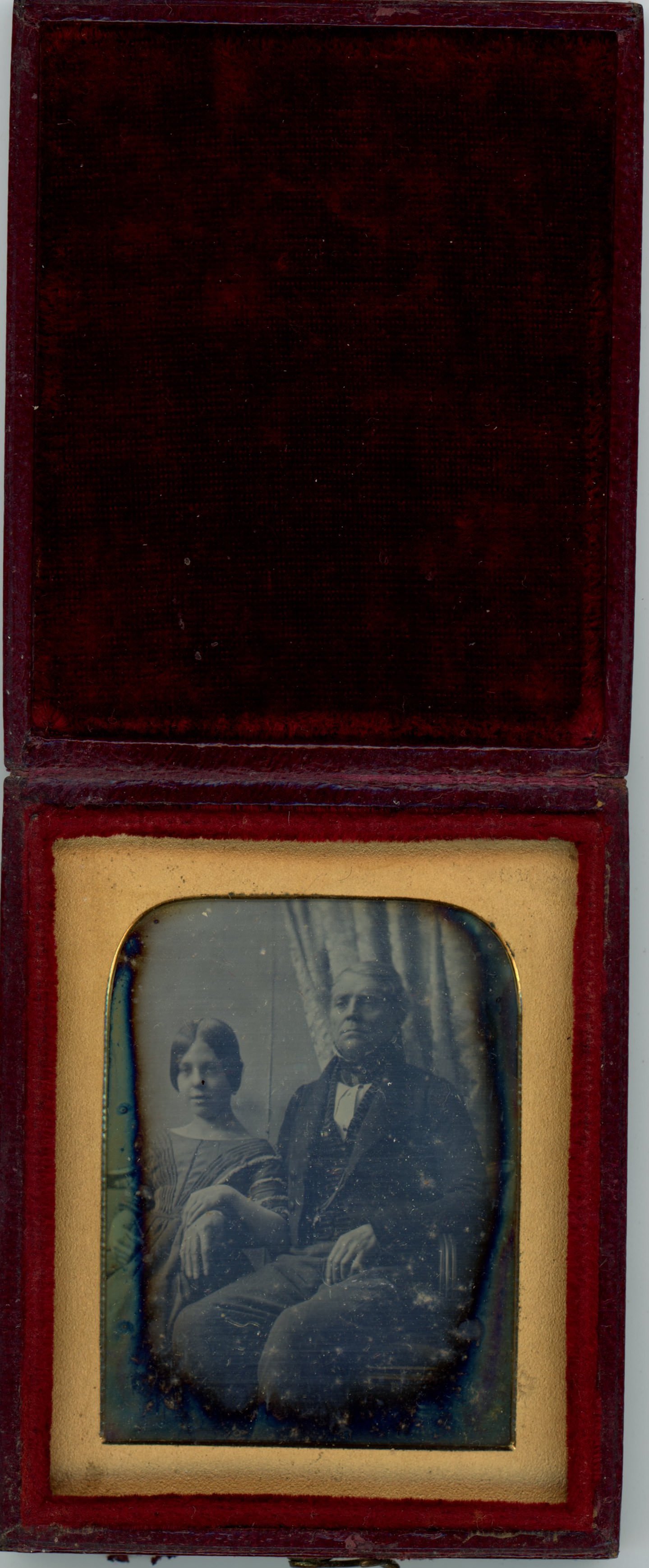 Daguerreotype in an open case. A young girl rests her elbow over the arm of a seated man. Both have pale skin, and are shown in front of a curtain. The rectangular gold mat has curved upper corners. The lid of the case is flipped up and the inside lined with dark red velvet.