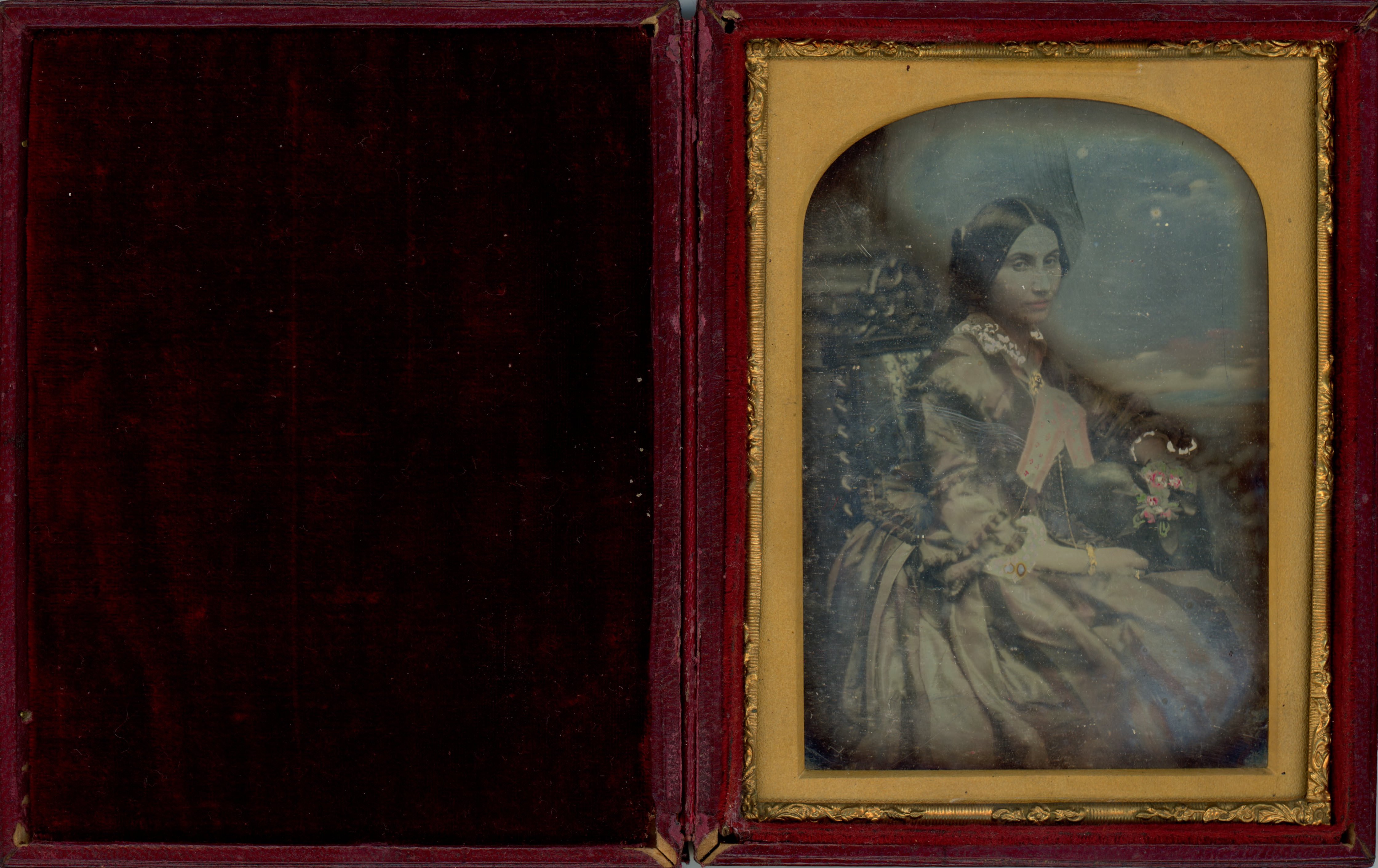 Daguerreotype in an open case. Shown from the knees up, a seated light-skinned woman rests one hand in voluminous folds of her full skirt. Her body angles to our right but she turns to look at us. The sky has been tinted blue and a few details pink.