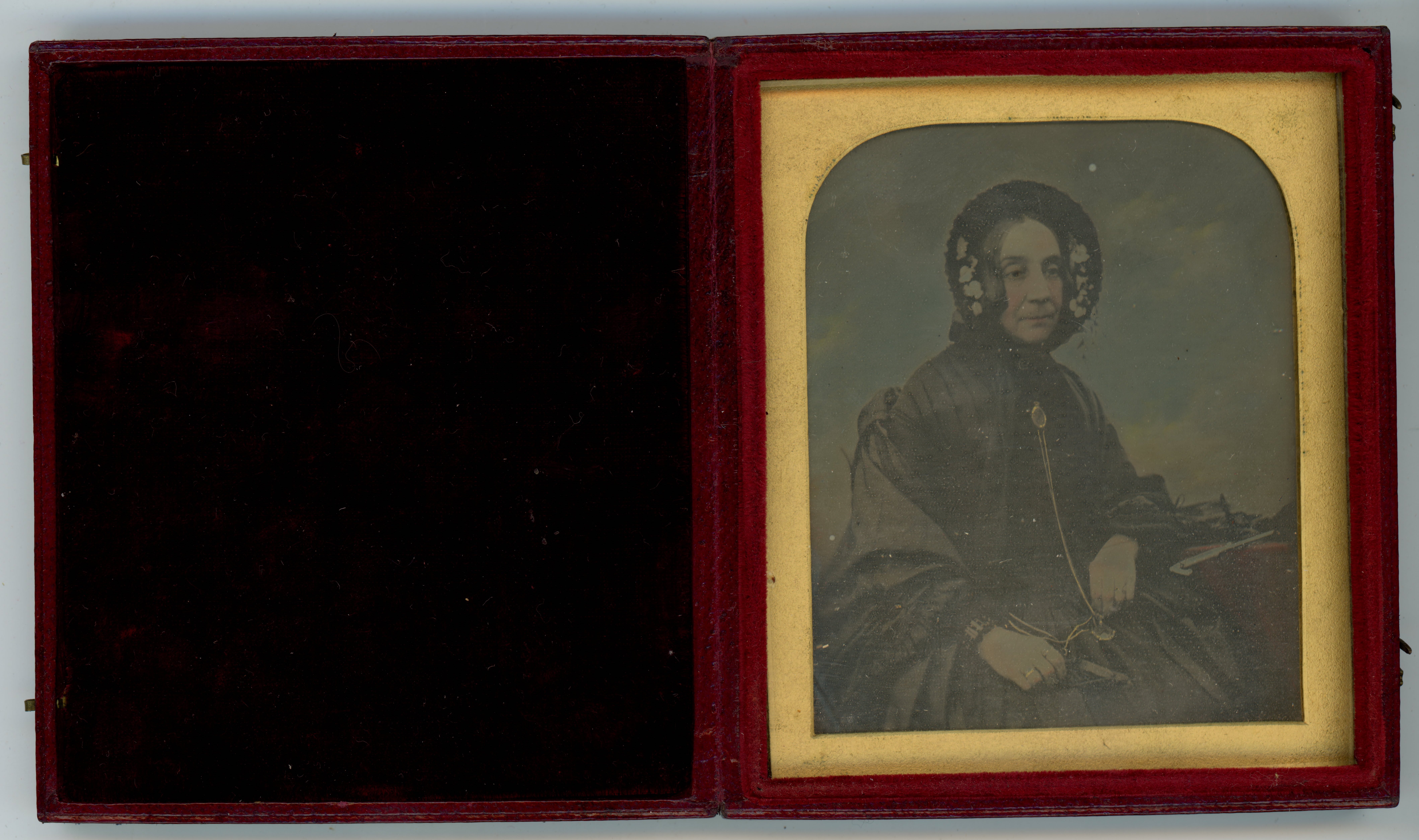 Daguerreotype in an open case. A pale-skinned woman wearing a bonnet and black dress has been hand-coloured to add rosy cheeks and a blue background. The photograph is in a gold frame in a red case, which opens like a book. The cover is lined with black fabric.