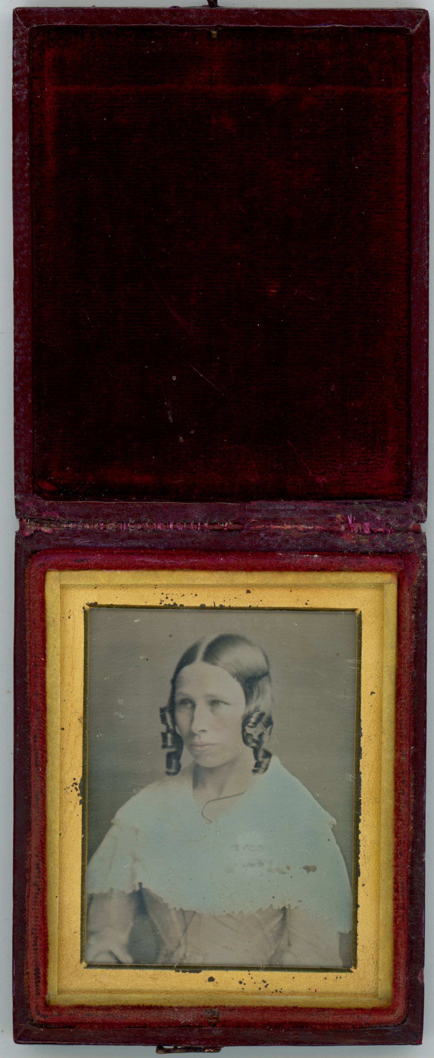 Daguerreotype in an open case. A light-skinned woman with curls framing her face looks off to our left. Pink has been added to her cheeks and dress beneath a white shawl. She is shown in a rectangular gold mat. The case flips up and the cover is lined with dark red fabric.