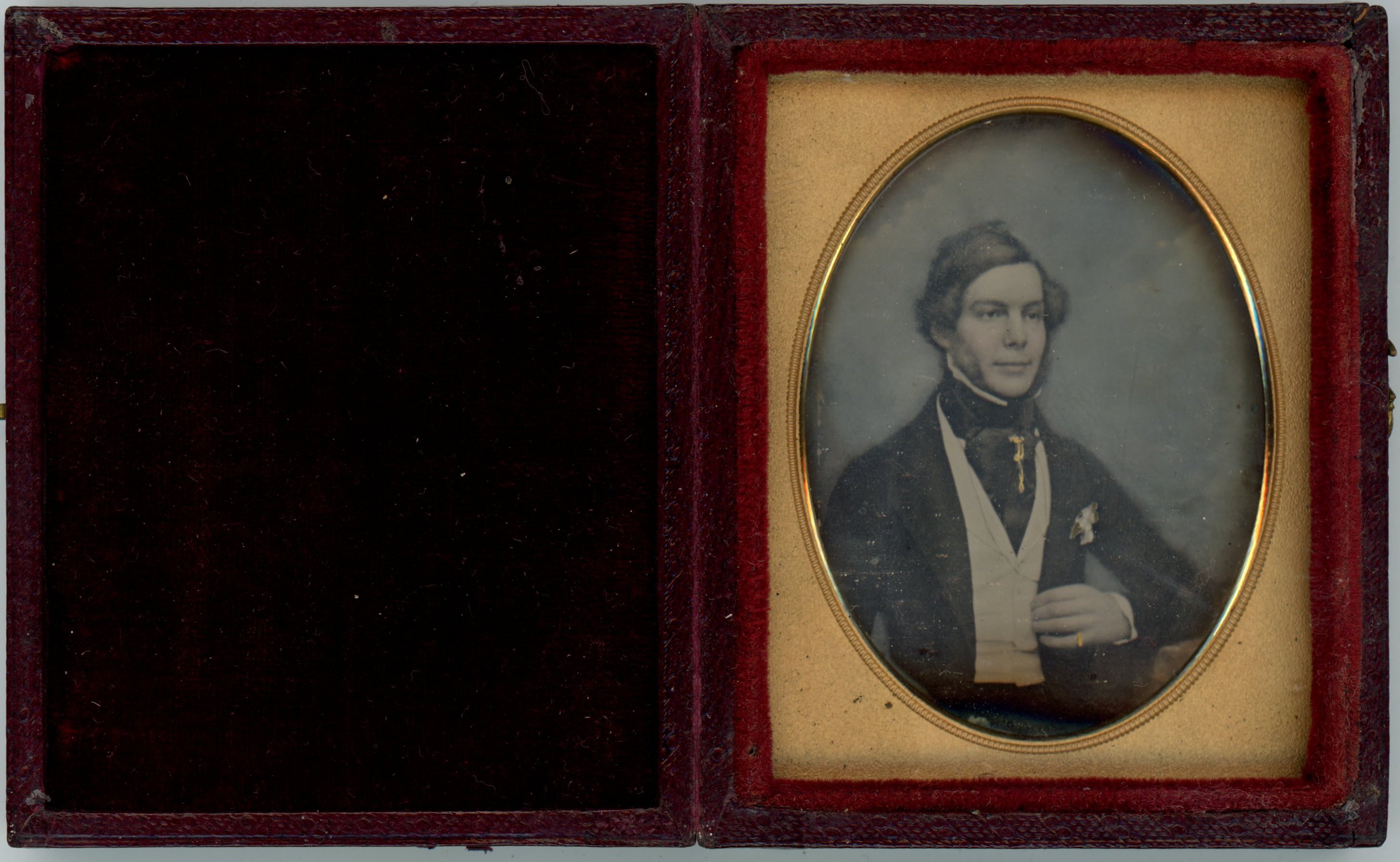 Daguerreotype in an open case. A pale-skinned man with mutton chops sits looking off to our right, a slight smile playing on his wide mouth. The background is tinted blue and his cheeks pink. A jewel on the puffy cravat at his throat may be coloured yellow.