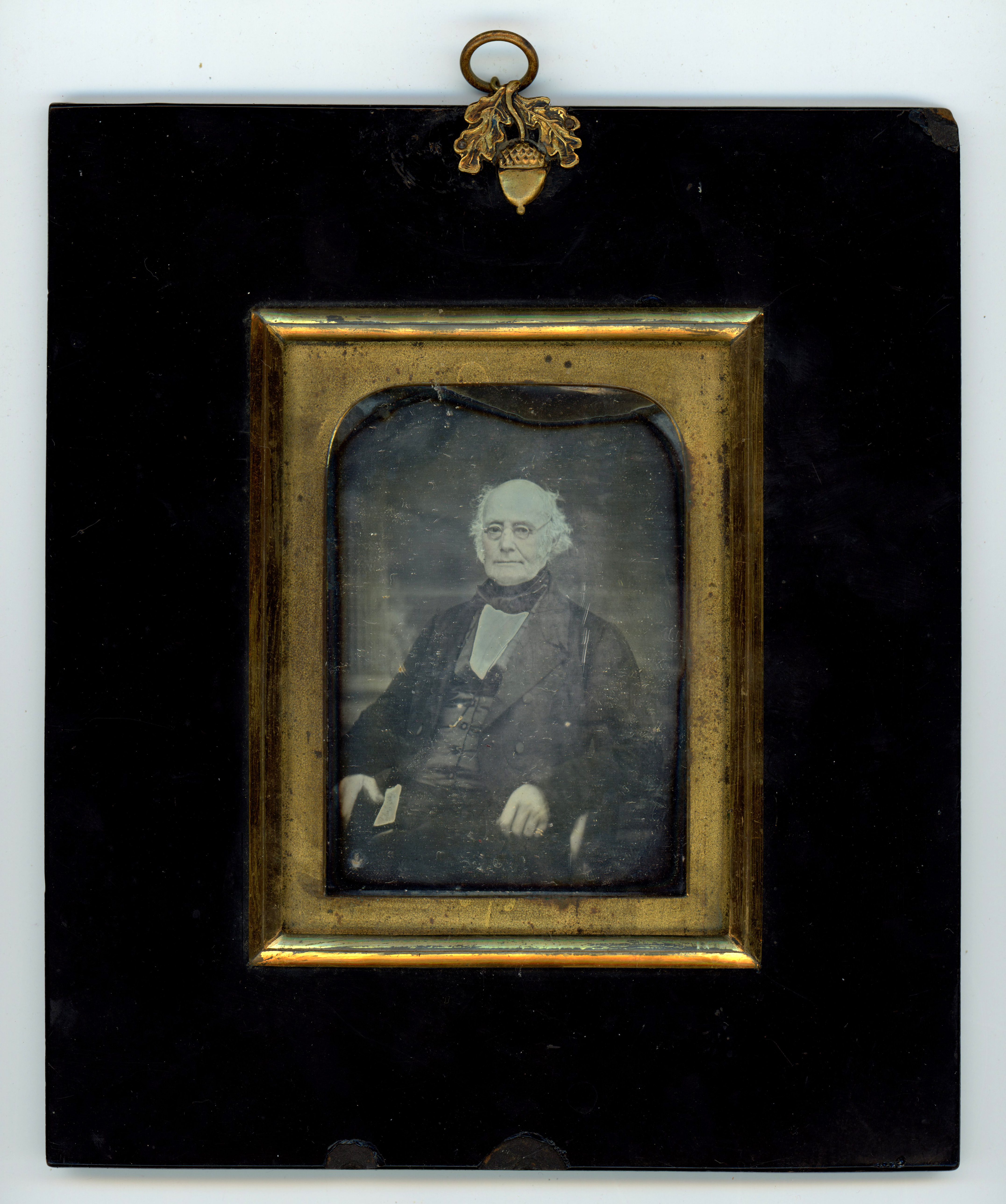 Daguerreotype. A pale-skinned, balding man with a deeply lined face sits looking off to our left. The rectangular gold mat is arched on the upper corners. A wide black frame surrounds it, and a gold acorn is at the top centre.