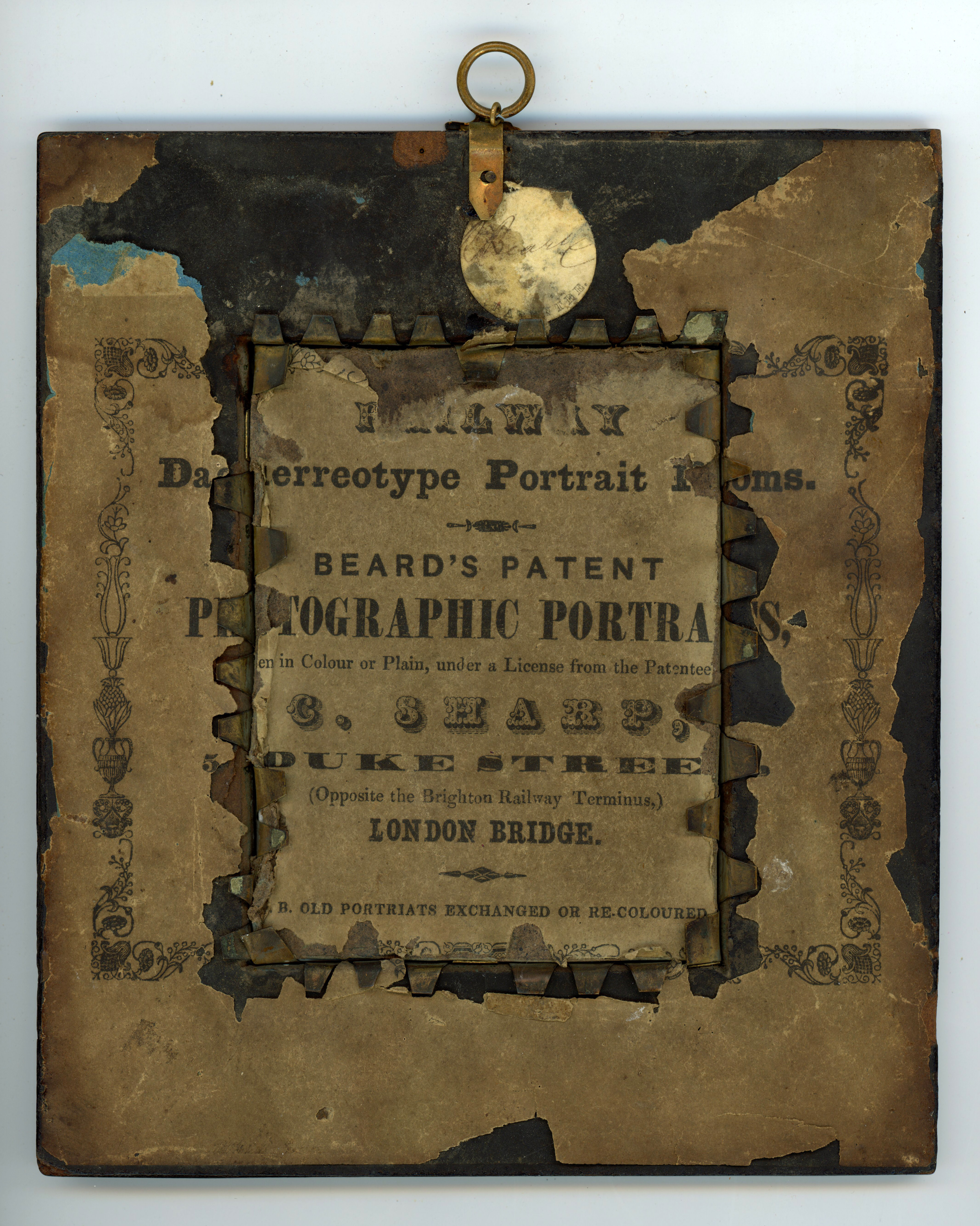 Label. A printed label is affixed to the back of a daguerreotype. The paper is chipped and discoloured, but some words and phrases can be made out. It reads as follows: “Daguerreotype Portrait Rooms Beard’s Patent Photograph Portraits...in Colour or Plain, under a License from the Patentee C. Sharp, Duke Street (Opposite the Brighton Railway Terminus,) London Bridge. Old Portraits Exchanged or Recoloured”.